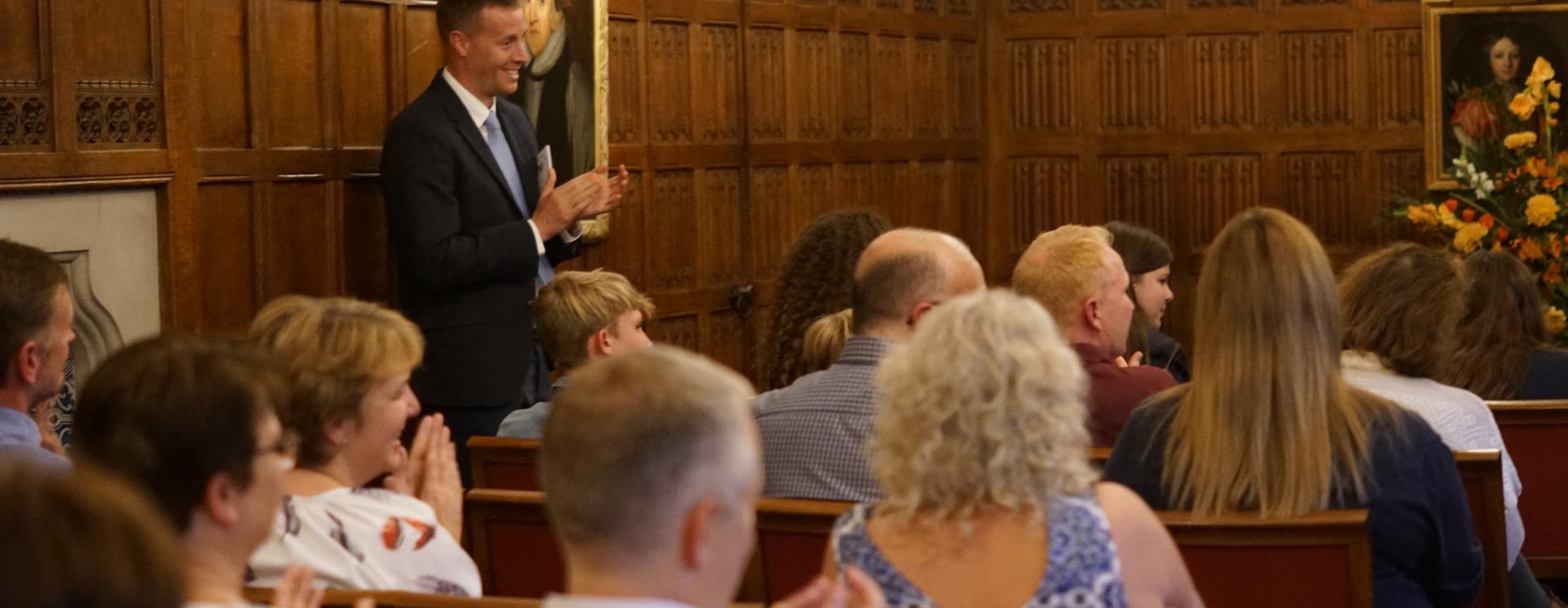 Parents and guardians attending an event at St Catharine's with their children. A male teacher is stood at the side of the seated audience, joining them in applause.